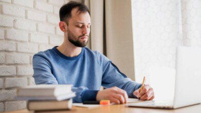Homem escrevendo em caderno enquanto estuda com notebook e livros sobre a mesa. Foto: Freepik