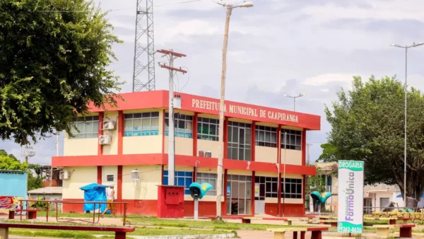 Prédio da Prefeitura Municipal de Caapiranga com dois pavimentos em tons de bege e detalhes em vermelho, apresentando grandes janelas de vidro e cercado por uma praça com bancos e árvores sob céu nublado. Foto: Divulgação/Prefeitura de Caapiranga-AM