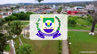 Vista aérea de uma praça arborizada e ruas comerciais da cidade de Alta Floresta D’Oeste, com o brasão oficial do município centralizado sobre a imagem.