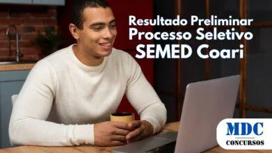 Homem jovem de pele morena e cabelo curto sentado à mesa segurando uma caneca e sorrindo enquanto olha para um notebook prateado em um ambiente aconchegante com cozinha de fundo - SEMED Coari (AM) divulga Resultado Preliminar