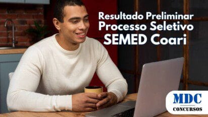 Homem jovem de pele morena e cabelo curto sentado à mesa segurando uma caneca e sorrindo enquanto olha para um notebook prateado em um ambiente aconchegante com cozinha de fundo - SEMED Coari (AM) divulga Resultado Preliminar