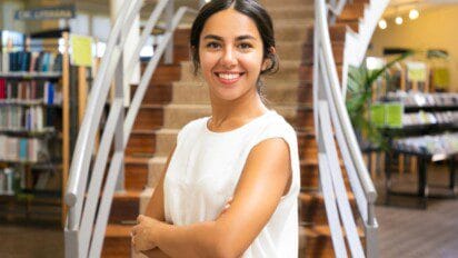 Mulher sorridente de braços cruzados em frente a escada em ambiente de biblioteca. Foto: pch.vector/Freepik