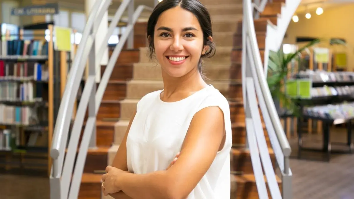 SEMED Juruti abre 20 vagas de até R$ 10.251,52 para Diretores Escolares 4 Mulher sorridente de braços cruzados em frente a escada em ambiente de biblioteca. Foto: pch.vector/Freepik