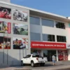 Fachada da Secretaria Municipal de Educação de Macapá, prédio moderno com painéis fotográficos sobre educação e inclusão, placa institucional em vermelho, veículos estacionados à frente e pessoas circulando na entrada. Foto: Divulgação/Prefeitura de Macapá