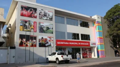 Fachada da Secretaria Municipal de Educação de Macapá, prédio moderno com painéis fotográficos sobre educação e inclusão, placa institucional em vermelho, veículos estacionados à frente e pessoas circulando na entrada. Foto: Divulgação/Prefeitura de Macapá