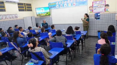 Professora dá aula para turma de alunos em sala de aula com carteiras azuis e quadro branco.