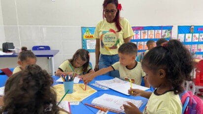 Professora acompanha crianças em sala de aula enquanto elas desenham e escrevem em cadernos sobre mesas azuis. Foto: Divulgação/Prefeitura de Marabá-PA