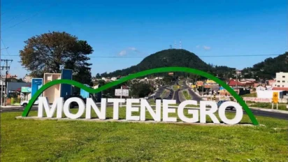Letreiro com o nome "Montenegro" em área urbana, com colina arborizada ao fundo e céu azul claro.