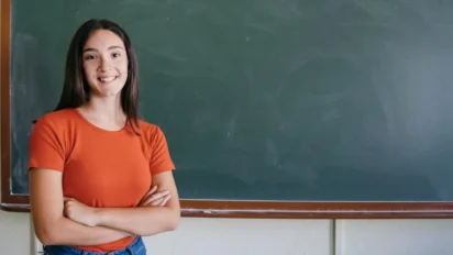 Mulher jovem de camiseta laranja sorrindo em frente a um quadro-negro. Foto: Freepik