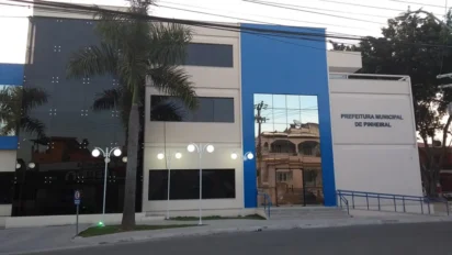 Fachada da Prefeitura Municipal de Pinheiral, prédio moderno em tons de branco, cinza e azul, com palmeira na frente e identificação institucional na parede lateral. Foto: Divulgação/Prefeitura de Pinheiral-RJ