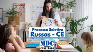 Imagem de uma sala de aula com uma professora segurando uma folha com um círculo colorido, aparentemente ensinando cores para crianças. Ao redor da mesa estão três crianças pequenas prestando atenção. O ambiente é colorido e decorado com plantas, brinquedos e cartazes educativos. No centro da imagem há um destaque de texto escrito “Processo Seletivo Russas - CE” e o logotipo “MDC Concursos”.