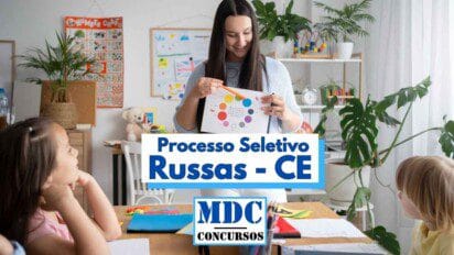 Imagem de uma sala de aula com uma professora segurando uma folha com um círculo colorido, aparentemente ensinando cores para crianças. Ao redor da mesa estão três crianças pequenas prestando atenção. O ambiente é colorido e decorado com plantas, brinquedos e cartazes educativos. No centro da imagem há um destaque de texto escrito “Processo Seletivo Russas - CE” e o logotipo “MDC Concursos”.