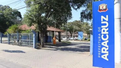 Entrada da Prefeitura de Aracruz com placa azul vertical, portaria ao fundo, árvores, bicicletas e carros estacionados.