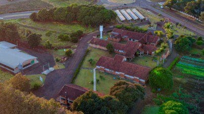 Vista aérea de unidade rural com prédios de telhado vermelho, áreas verdes, estufas agrícolas e vias internas asfaltadas.