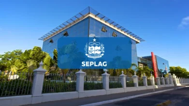 Fachada do prédio da SEPLAG AL com estrutura de vidro espelhado sob céu azul, cercada por gradil branco e vegetação, apresentando o logotipo centralizado em um retângulo azul sobreposto à imagem. Foto: Divulgação/SEPLAG-AL