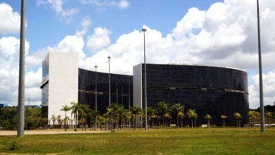 Vista do edifício moderno da Cidade Administrativa de Minas Gerais, com fachada espelhada, palmeiras ao redor e céu parcialmente nublado ao fundo.