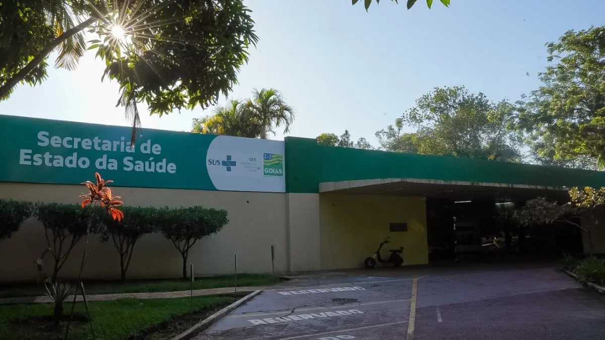 Fachada da Secretaria de Estado da Saúde de Goiás (SES-GO) com letreiro sobre muro verde e branco, cercada por árvores sob a luz do sol.