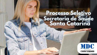 Mulher loira com jaqueta jeans e camiseta branca trabalha em um laptop prateado em uma mesa externa de café. Ela está concentrada na tela, com a mão ajustando a posição do dispositivo. Seu reflexo aparece na janela atrás dela, e há um cinzeiro sobre a mesa - SES SC abre 33 Vagas com salários de até R$ 10.243 em Processo Seletivo