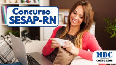 Mulher jovem com cabelos longos e castanhos sorrindo enquanto usa um smartphone sentada em um sofá. Ela veste uma blusa preta com um cardigã rosa e tem um laptop no colo. O ambiente é aconchegante, com uma estante branca ao fundo cheia de livros, decorações e plantas - SESAP RN abre 517 vagas com salários de até R$ 4.270 na Área da Saúde