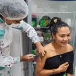 Profissional de saúde aplica vacina no braço de uma jovem sorridente em ambiente clínico, com armário de medicamentos ao fundo.