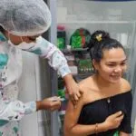 Profissional de saúde aplica vacina no braço de uma jovem sorridente em ambiente clínico, com armário de medicamentos ao fundo.