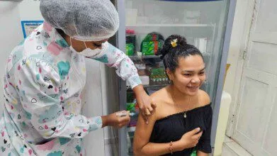 Profissional de saúde aplica vacina no braço de uma jovem sorridente em ambiente clínico, com armário de medicamentos ao fundo.
