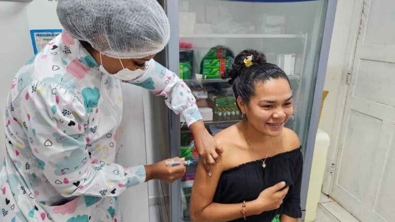 Profissional de saúde aplica vacina no braço de uma jovem sorridente em ambiente clínico, com armário de medicamentos ao fundo.
