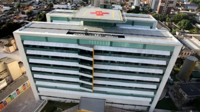 Vista aérea do hospital Santa Casa do Pará em Belém, com heliponto no telhado exibindo o símbolo da cruz vermelha, e fachada modernista de vidro e metal cercada por outros edifícios urbanos.
