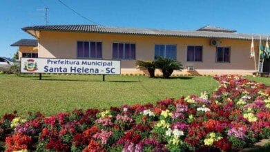 Prédio da Prefeitura Municipal de Santa Helena-SC com jardim florido à frente, placa com o nome do município e bandeiras ao lado.