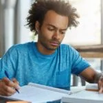 Homem jovem estudando em uma mesa com caderno e livro, concentrado na leitura e escrita. Foto: wayhomestudio/Freepik