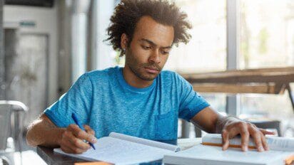 Homem jovem estudando em uma mesa com caderno e livro, concentrado na leitura e escrita. Foto: wayhomestudio/Freepik