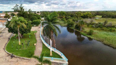 Processos Seletivos 2 Vista aérea de parque urbano com equipamentos de ginástica, palmeiras e calçadão à margem de um rio em Santa Rita de Cássia, BA