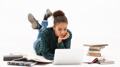 Jovem deitada no chão estudando em frente a um notebook, cercada por livros abertos e empilhados. Foto: cookie_studio/Freepik