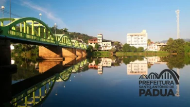 Ponte verde sobre o rio em Santo Antônio de Pádua, com reflexo na água, prédios ao fundo, céu azul e marca da Prefeitura do município no canto da imagem. Foto: Divulgação