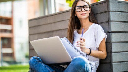 Jovem sentada ao ar livre com notebook e caderno no colo, segurando caneta e olhando pensativa para o lado. Foto: diana.grytsku/Freepik