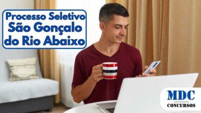 Homem sentado à mesa usando celular, segurando uma caneca listrada e com um notebook à sua frente, em ambiente doméstico com sofá e cortinas ao fundo - São Gonçalo do Rio Abaixo (MG) abre 34 Vagas com salário de R$ 3 mil em Processo Seletivo