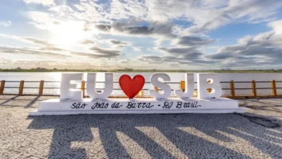 Estrutura com a frase “EU ❤️ SJB” em frente ao rio, ao pôr do sol, com base escrita “São João da Barra – RJ Brasil” e céu com nuvens ao fundo. Foto: Divulgação/Prefeitura de São João da Barra-RJ