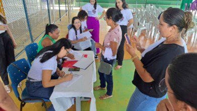 Processos Seletivos 2 Grupo de mulheres participa de atendimento em uma mesa com formulários, em ambiente coberto com cadeiras plásticas ao fundo.