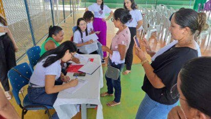 Grupo de mulheres participa de atendimento em uma mesa com formulários, em ambiente coberto com cadeiras plásticas ao fundo.