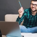 Homem sorridente com óculos sentado em sofá, segurando um lápis e usando notebook, com papéis sobre a mesa.
