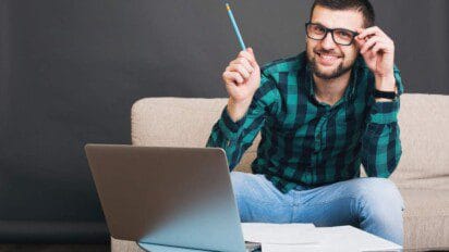 Homem sorridente com óculos sentado em sofá, segurando um lápis e usando notebook, com papéis sobre a mesa.