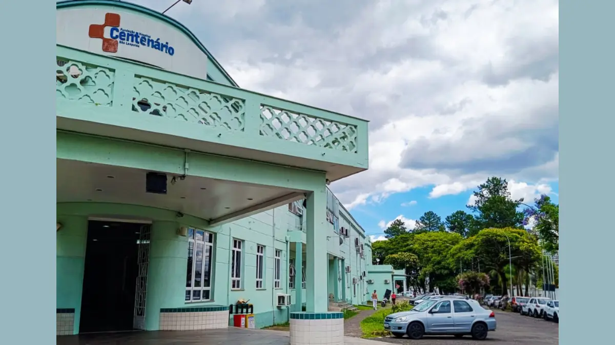 São Leopoldo/RS: FHC abre concurso com 191 vagas e salários de até R$ 11 mil 1 Fachada da Fundação Hospital Centenário em São Leopoldo - FHC, apresentando um edifício de cor verde clara com arquitetura clássica, letreiro azul no topo e estacionamento com carros ao lado sob céu nublado.