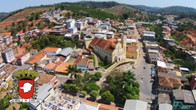 Vista aérea do centro de São Miguel do Anta com prédios residenciais, ruas, áreas verdes e igreja em destaque, com morros ao fundo e brasão do município no canto da imagem. Foto: Divulgação/Prefeitura de São Miguel do Anta-MG