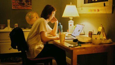 Pessoa sentada em mesa de estudo à noite, concentrada em notebook, com ambiente iluminado por abajur e itens como copos, latas e cadernos ao redor.