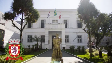 Fachada do prédio histórico da Prefeitura de São Vicente, com estátua ao centro, jardim frontal, árvores laterais e bandeiras do Brasil, de São Paulo e do município no topo. Foto: Divulgação/Prefeitura de São Vicente-SP