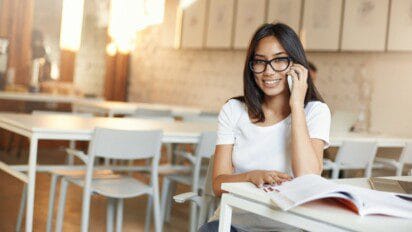 Mulher jovem com óculos sorrindo, sentada em sala de aula, falando ao celular e com livro aberto sobre a mesa. Foto: lookstudio/Freepik