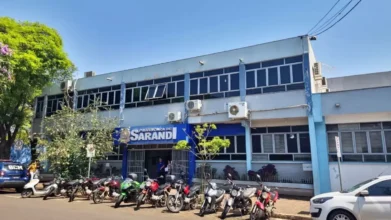 Fachada da Prefeitura de Sarandi em um dia ensolarado, apresentando estrutura de dois andares com janelas de vidro e letreiro azul na entrada, além de diversas motocicletas estacionadas em frente. Foto: Divulgação/Prefeitura de Sarandi-PR