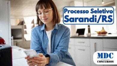 Mulher jovem de óculos olhando para o celular enquanto está apoiada em uma bancada de cozinha moderna e iluminada - Sarandi RS lança Processo Seletivo 2025 para Agentes de Saúde