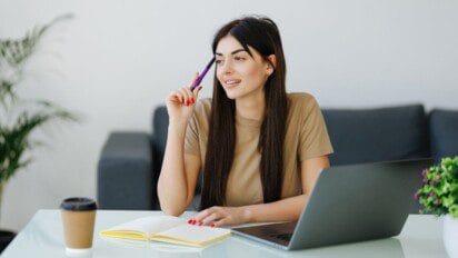 Mulher sentada à mesa com laptop, caderno e café, segurando caneta e sorrindo pensativa. Foto: Divulgação/Araripe-CE