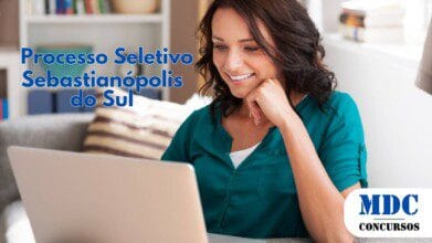 Processos Seletivos 6 Mulher jovem de cabelos castanhos e ondulados, usando uma blusa verde, sorrindo enquanto olha para a tela de um laptop prateado em um ambiente aconchegante com sofá, almofadas e estante ao fundo - Sebastianópolis do Sul (SP) lança Processo Seletivo